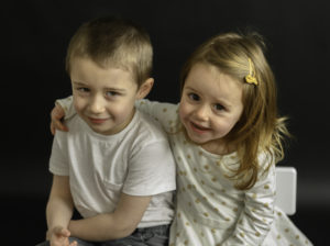 Un frère et sa soeur assis sur un banc, la petite fille tient son grand frère par l'épaule, les deux sourient, photographe d'enfants Houilles