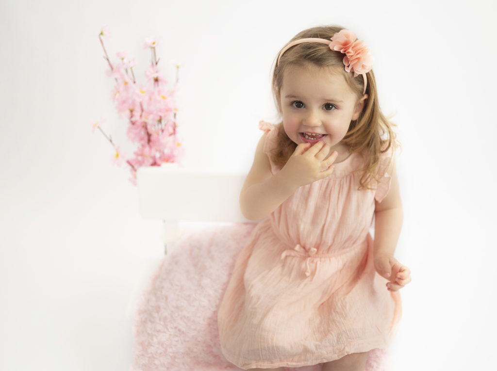 Petite fille en robe rose et bandeau rose fleuri, assise sur un banc auquel des fleurs roses sont accrochées Photographe enfants Houilles