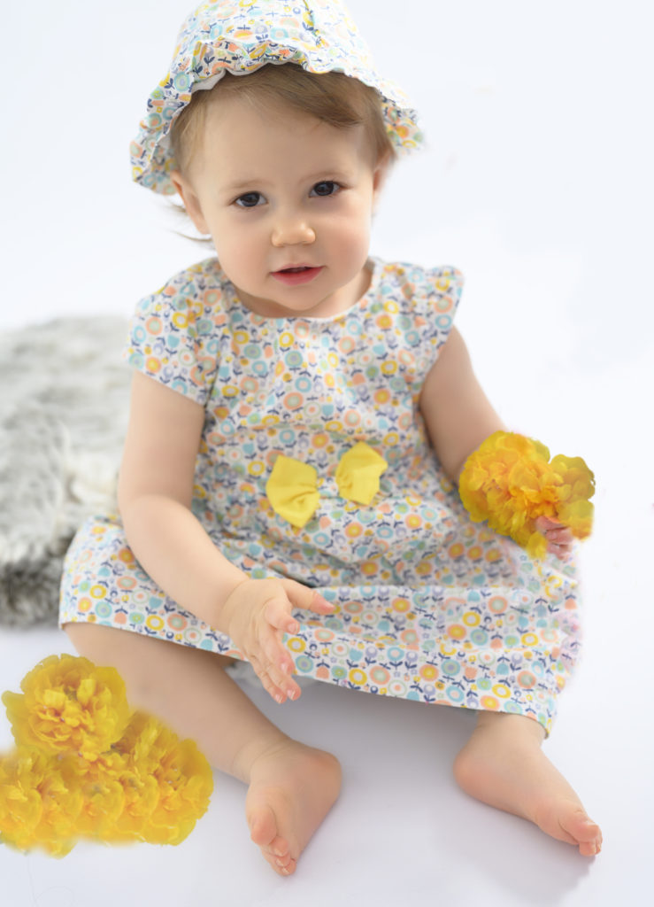 Petite fille en robe fleurie jaune assise sur une fourrure, joue avec des fleurs Photographe bébé Houilles Photographe Houilles