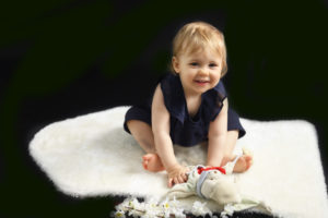 Bébé fille assise sur un tapis blanc, faisant un grand sourire avec doudou dans les mains, photographie de bébé Houilles
