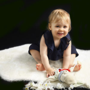 Bébé fille assise sur un tapis blanc, faisant un grand sourire avec doudou dans les mains, photographie de bébé Houilles