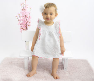 Bébé de 10 mois assise sur un petit banc, robe blanche bandeau blanc fleurs et tapis roses Photographe bébé Houilles photographe Houilles