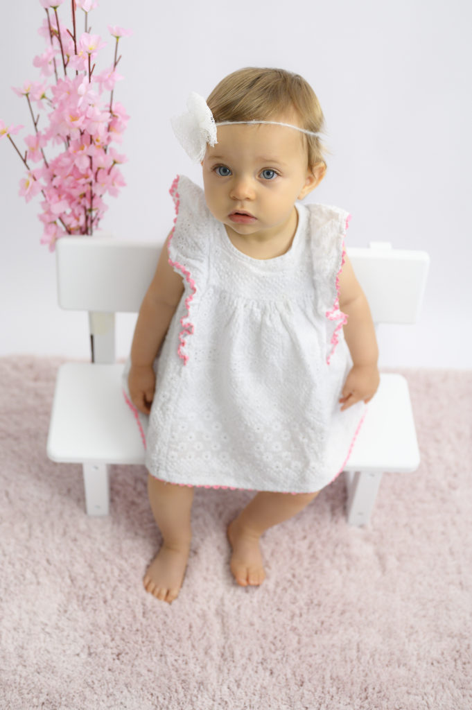 Bébé de 10 mois assise sur un petit banc, robe blanche bandeau blanc fleurs et tapis roses Photographe bébé Houilles photographe Houilles