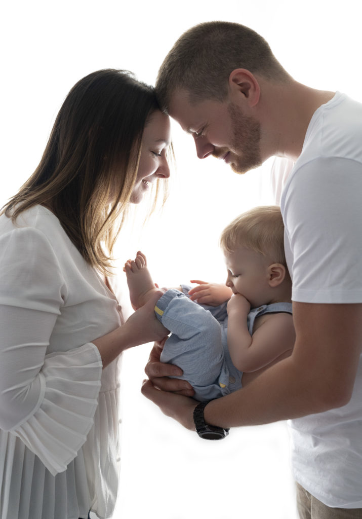 Image en contrejour de bébé de huit mois dans les bras de ses parents, de profil, front contre front. photographe bébé famille Houilles Yvelines la Défense