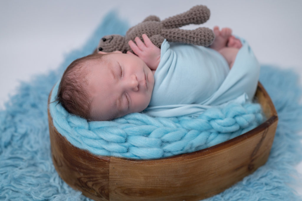 nouveau-né emmailloté de bleu endormi dans un coeur en bois sur une tresse en laine bleue tenant un petit nounours dans ses mains photographe naissance nouveau-né Houilles Yvelines la Défense