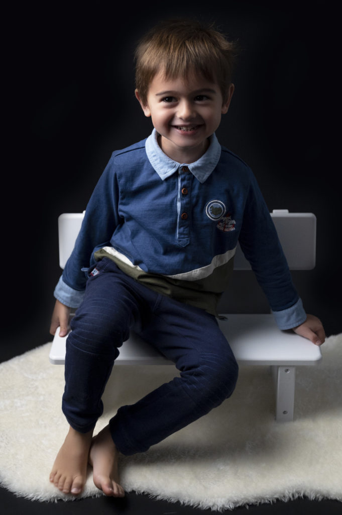 portrait d'un petit garçon qui sourit assis sur un petit banc blanc. Photographe bébé enfant famille Houilles Yvelines la Défense