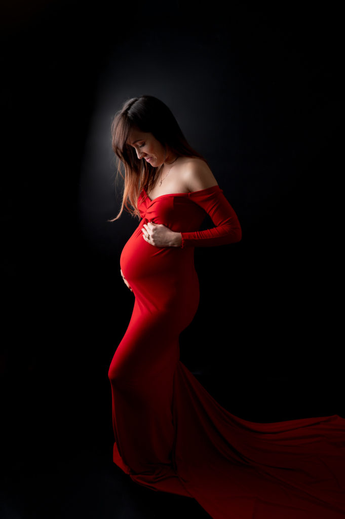 femme enceinte en robe rouge à traine sur fond noir regarde son ventre mains sur le ventre de la femme enceinte photographe grossesse maternité Houilles Yvelines la Défense
