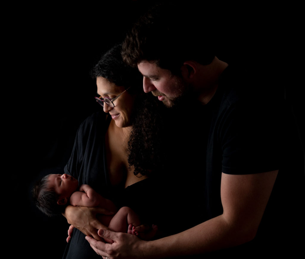 Jeunes parents sur fond noir tenant leur nouveau-né dans leurs bras photographe naissance famille Houilles Yvelines la Défense