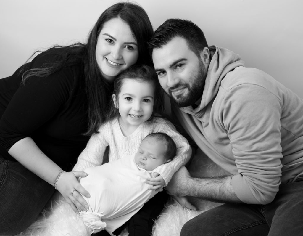 portrait noir et blanc d'une famille avec la grande soeur qui tient son tout-petit frère emmailloté dans ses bras photographe naissance famille Houilles la Défense