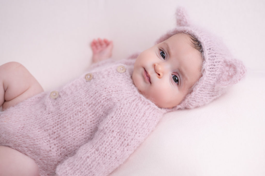 Bébé de quatre mois en body rose et bonnet rose à oreilles d'ours en laine doudou sourit, photographe bébé famille Houilles Yvelines la Défense