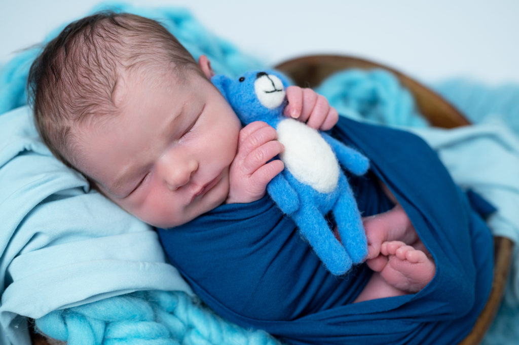 Nouveau-né emmailloté de bleu pétrole tenant dans ses mains un petit nounours bleu photographe naissance nouveau-né Houilles Yvelines la Défense
