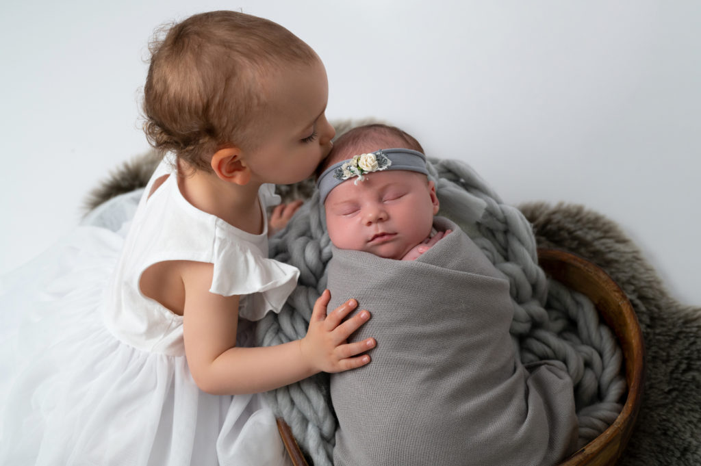 bébé de deux ans faisant un bisou à sa petite soeur nouvelle née emmaillotée de gris endormie dans un coeur en bois photographe naissance nouveau-né Houilles Yvelines la Défense