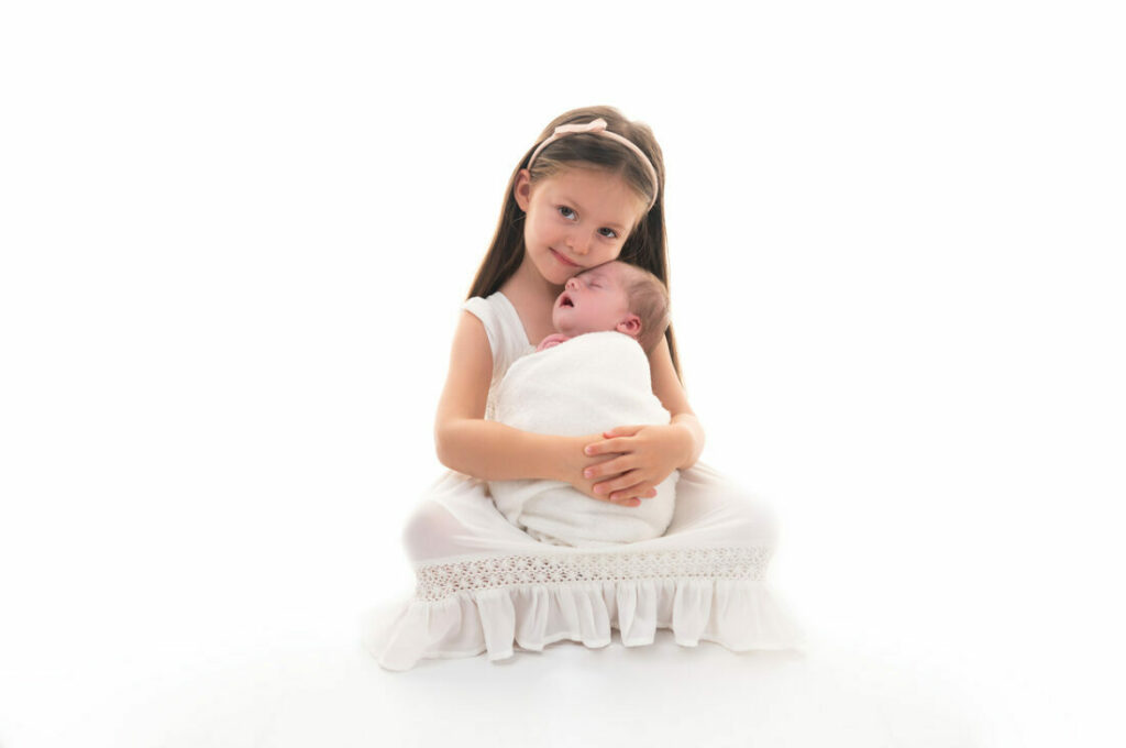 grande sœur qui tient sa petite sœur dans les bras fond blanc photographe naissance Houilles photographe naissance la Défense photographe Houilles photographe Versailles photographe Rueil-Malmaison photographe Chatou photographe Yvelines photographe Ile-de-France