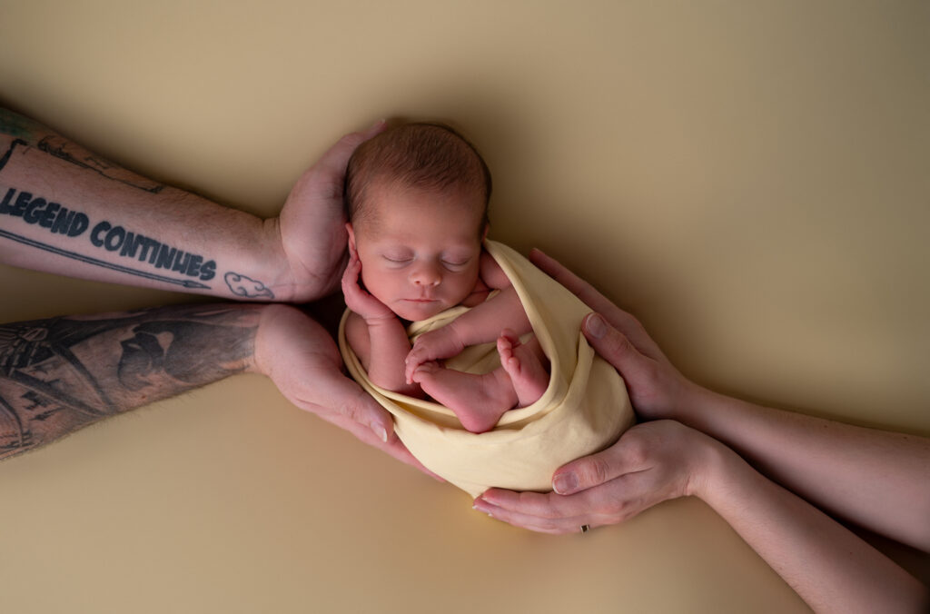 astuces pour apaiser bébé nouveau-né emmaillotée de jaune endormie dans les mains de ses parents photographe naissance Houilles photographe naissance Yvelines photographe naissance Versailles photographe naissance Ile de France photographe naissance Carrières sur Seine photographe naissance Chatou photographe naissance Paris